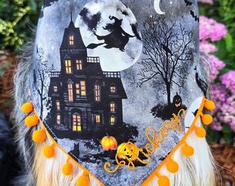 May include: A triangular pet bandana featuring a gray and black Halloween scene with a haunted house, a witch flying across a full moon, bats, and trees. The fabric is trimmed with orange pom-poms and embroidered with the name Hickory in orange.