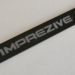May include: A black rectangular plate with the word "IMPREZIVE" in grey lettering. The plate is accompanied by two screws and two cylindrical fasteners. The items are on a light grey surface.