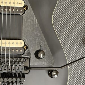Puede incluir: Una guitarra eléctrica negra con una golpeador negro y herrajes cromados. La guitarra tiene una sola pastilla humbucker y un puente trémolo.