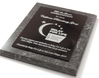 May include: A black and silver plaque with the text "With Our Greatest Appreciation We Thank California Philanthropy Group" and the logo for Relay For Life, American Cancer Society. The plaque also includes the text "For your support and dedication to Relay For Life in San Luis Obispo. Your hard work does not go unnoticed. Presented by Relay For Life in San Luis Obispo 2019."