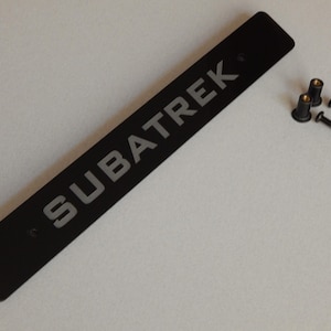 Puede incluir: Accesorio de coche rectangular negro con la palabra "SUBATREK" en letras grises. Incluye dos tornillos y dos remaches de montaje. El accesorio es probablemente para un vehículo.