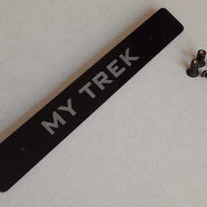 May include: A black rectangular plate with the text "MY TREK" in silver lettering. Two black screws and two black fasteners are to the right of the plate. The plate has two holes for mounting.