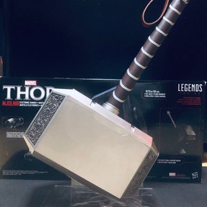 May include: A silver and black Thor's hammer replica with a brown leather strap. The hammer is on a clear plastic stand and has intricate details. The box behind the hammer says "Marvel Legends Series" and "Thor Mjolnir Electronic Hammer".