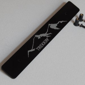 May include: Black metal plate with a mountain range design and the word "TREKKIN'" engraved in silver. The plate comes with two black screws and two black nuts.