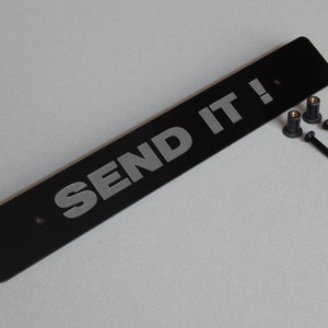 May include: A black rectangular plate with the words "SEND IT!" in silver lettering. Two black screws and two brass-colored inserts are included. The plate has two pre-drilled holes for mounting.