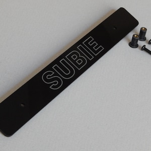 以下が含まれることがあります： 黒い金属製のナンバープレートフレームに、白い文字で「SUBIE」と刻印されています。フレームには、黒いネジとナットが4つ付属しています。