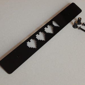 Puede incluir: Placa de metal negro con cuatro recortes de corazón pixelados. La placa viene con tres tornillos negros y una tuerca dorada.