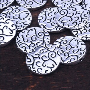 May include: A collection of silver metal round beads with a decorative swirl pattern. The beads are scattered on a dark brown wooden surface.