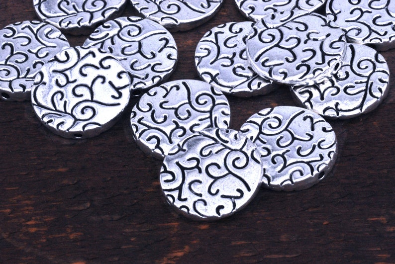 May include: A collection of silver metal round beads with a decorative swirl pattern. The beads are scattered on a dark brown wooden surface.