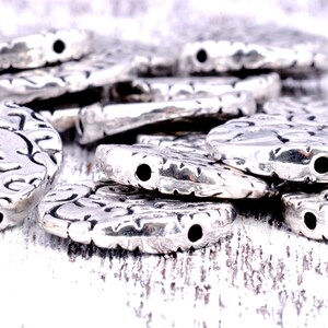 May include: A pile of silver-toned metal beads with a decorative swirl pattern. The beads have a hole in the center for stringing.