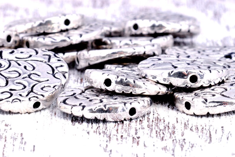 May include: A pile of silver-toned metal beads with a decorative swirl pattern. The beads have a hole in the center for stringing.