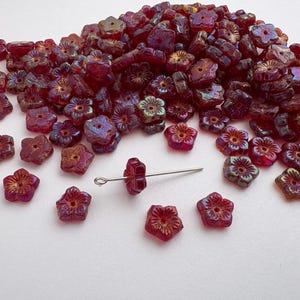 May include: A large collection of small, translucent, deep red flower-shaped beads with an iridescent finish. Each bead features a central hole for stringing. One bead is threaded onto a thin, silver-colored metal headpin with a looped end.