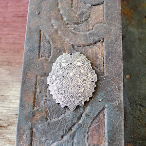 May include: Silver-toned metal charm with an intricate floral design. The charm is oval-shaped and has a detailed pattern of leaves and flowers.