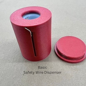 Può includere: Un erogatore di filo di sicurezza in plastica rossa con un tagliafilo in metallo. L'erogatore ha un coperchio e una fessura per il passaggio del filo. Il testo "Basic Safety Wire Dispenser" è stampato sulla superficie sotto l'erogatore.