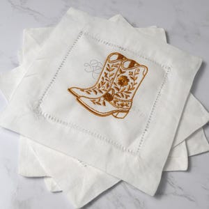 May include: White square linen cocktail napkins with an embroidered design of a cowboy boot in shades of gold and brown. The boot features floral and leaf details. The napkins are stacked, showcasing the embroidered design.