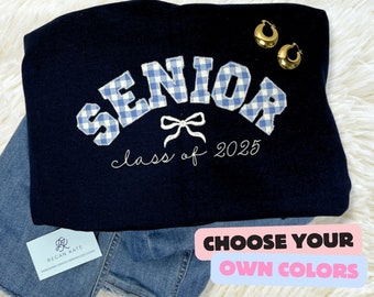 May include: A navy blue sweatshirt with the word "SENIOR" in white letters with a blue and white gingham pattern. The letters are arched and have a white bow underneath. The text "class of 2025" is written in white cursive below the word "SENIOR".