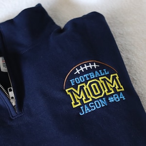 May include: Navy blue quarter-zip sweatshirt with an embroidered design that says "Football Mom Jason #84" in yellow and blue lettering.