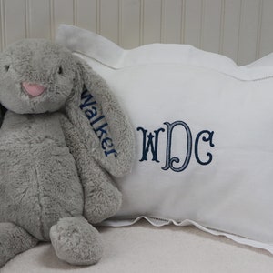 May include: A white pillow with a blue embroidered monogram "WDC" and the name "Walker" embroidered on the side. A gray plush bunny sits on the pillow.
