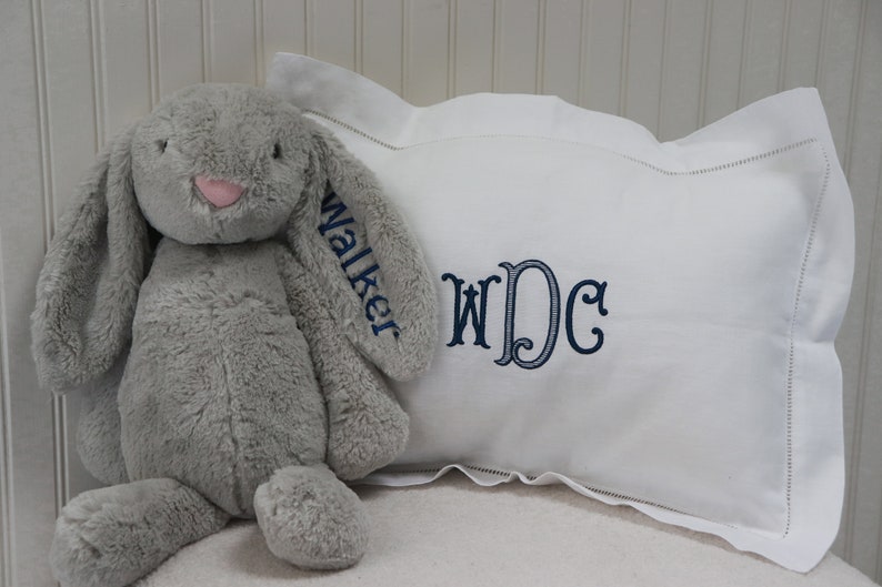 May include: A white pillow with a blue embroidered monogram "WDC" and the name "Walker" embroidered on the side. A gray plush bunny sits on the pillow.