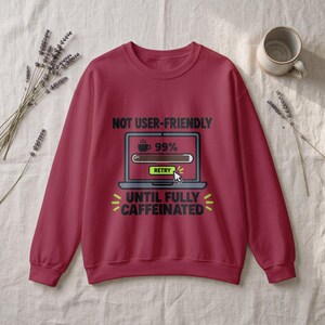 Puede incluir: Una sudadera de cuello redondo color rojo oscuro con un gráfico de una pantalla de portátil con una barra de carga al 99 por ciento, un icono de taza de café y un botón de retry. El texto dice NOT USER-FRIENDLY UNTIL FULLY CAFFEINATED.