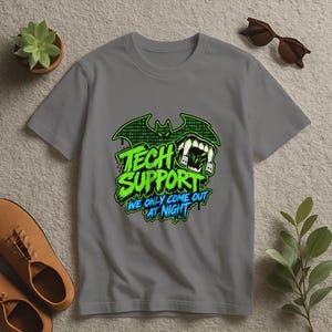 May include: A grey short-sleeved t-shirt featuring a graphic of a bat silhouette filled with binary code, a pair of fangs, and the text "TECH SUPPORT WE ONLY COME OUT AT NIGHT" in bright green and blue lettering on the front chest area.