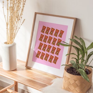 May include: A framed print with a pink background and a repeating pattern of brown and orange snack bags. The print is hanging on a white wall above a wooden table.