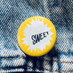 May include: A round, yellow button with the word "SWEET" in black, stylized text. The button has a white burst design around the edges and a light blue shape behind the word. The button is pinned to a blue denim jacket.