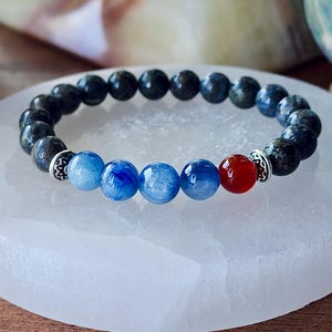 May include: A blue and black gemstone bracelet with a single red gemstone bead. The bracelet is on a white selenite charging plate.