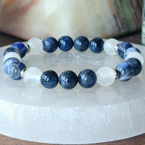 May include: A beaded bracelet featuring alternating dark blue and white spherical beads. The bracelet is strung on a stretchy cord and has silver-colored metal accents. The beads are smooth and polished, creating a striking contrast.