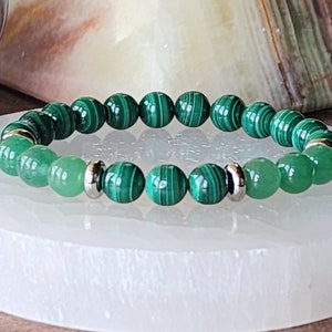 May include: A green malachite and green aventurine beaded bracelet with silver spacers. The bracelet is on a white selenite charging plate.