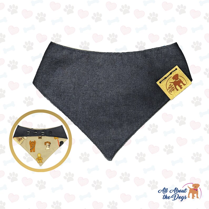K&ouml;nnte beinhalten: Ein dunkelgraues Denim-Hundehalstuch mit einem braunen und gelben Etikett mit der Aufschrift "All About the Dogs". Das Halstuch ist reversibel und hat eine beige Seite mit einem Muster von Hunden in verschiedenen Posen.