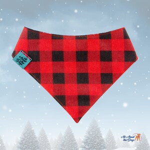 May include: Red and black buffalo plaid dog bandana with a turquoise tag that reads "All About the Dogs". The bandana is triangular and has a stitched edge. The background is a winter scene with falling snow and evergreen trees.