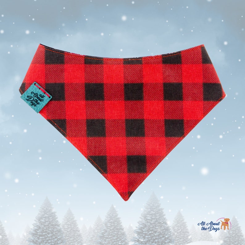 May include: Red and black buffalo plaid dog bandana with a turquoise tag that reads "All About the Dogs". The bandana is triangular and has a stitched edge. The background is a winter scene with falling snow and evergreen trees.