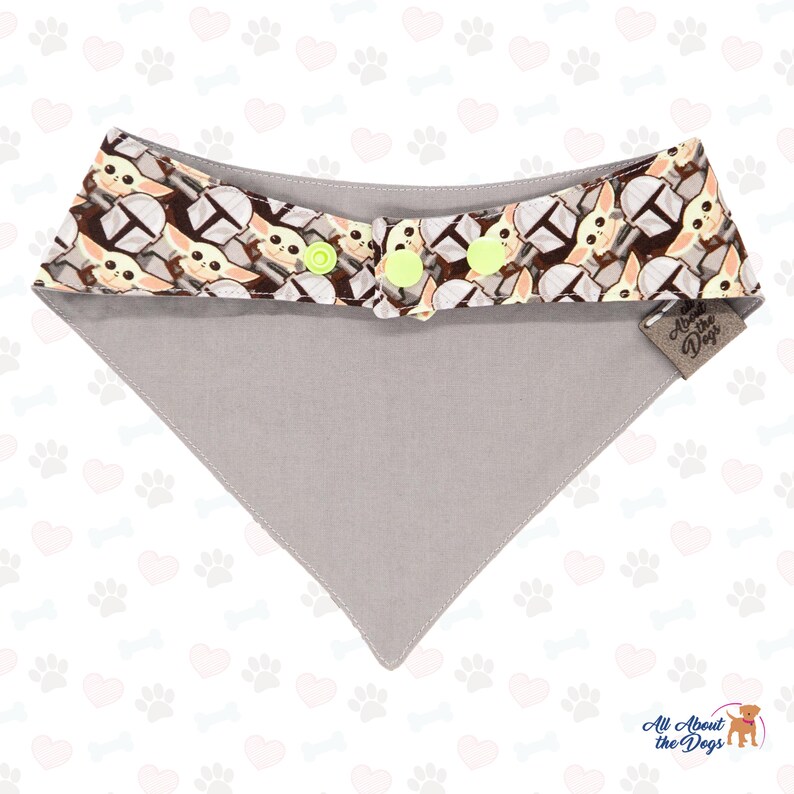 May include: A grey dog bandana with a brown and yellow pattern of a popular character from a science fiction film. The bandana has two yellow buttons and a brown tag that says "All About the Dogs".