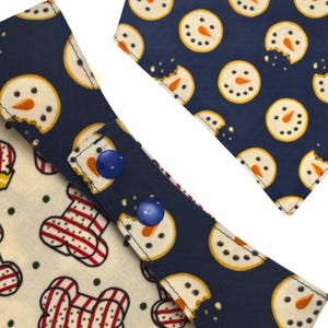 May include: Close-up of two dog bandanas with a festive winter theme. One bandana features a navy blue background with snowman cookie designs, while the other has a bone and ornament pattern. Both have blue snap closures.
