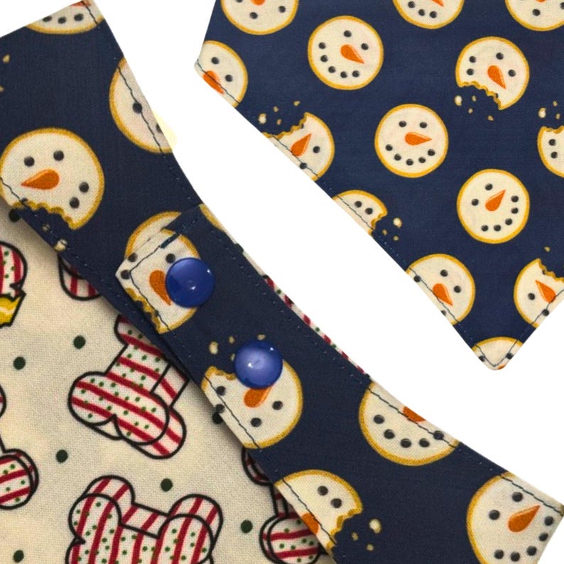 May include: Close-up of two dog bandanas with a festive winter theme. One bandana features a navy blue background with snowman cookie designs, while the other has a bone and ornament pattern. Both have blue snap closures.