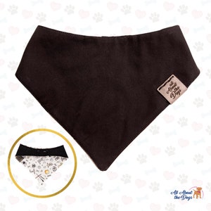 Op de afbeelding: Een bruine hondenbandana met een omkeerbaar ontwerp. De ene kant is effen bruin en de andere kant heeft een witte achtergrond met een zwart-wit patroon van honden gerelateerde pictogrammen. De bandana heeft een bruin lederen label met de tekst "All About the Dogs" in wit.
