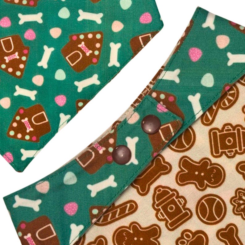 K&ouml;nnte beinhalten: Hunde-Bandana mit gr&uuml;nem Hintergrund und einem Muster aus wei&szlig;en Knochen, rosa Formen und Lebkuchen-Hundekeksen. Das Bandana hat einen Druckknopfverschluss mit zwei braunen Druckkn&ouml;pfen.