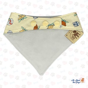May include: A light grey and yellow dog bandana with a pattern of small green and brown characters. The bandana has three yellow buttons and a brown tag that says "All About the Dogs".