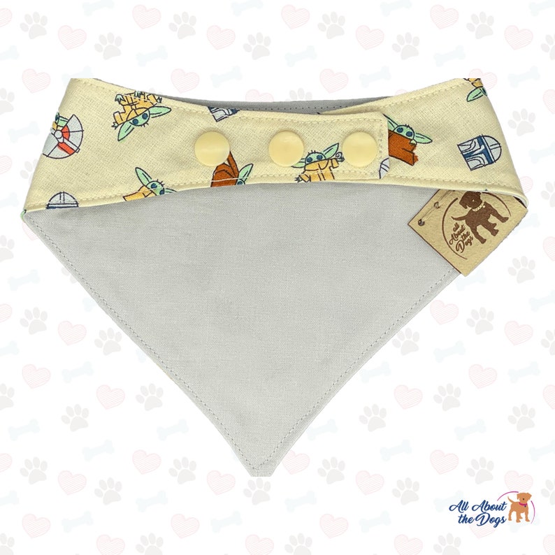 May include: A light grey and yellow dog bandana with a pattern of small green and brown characters. The bandana has three yellow buttons and a brown tag that says "All About the Dogs".