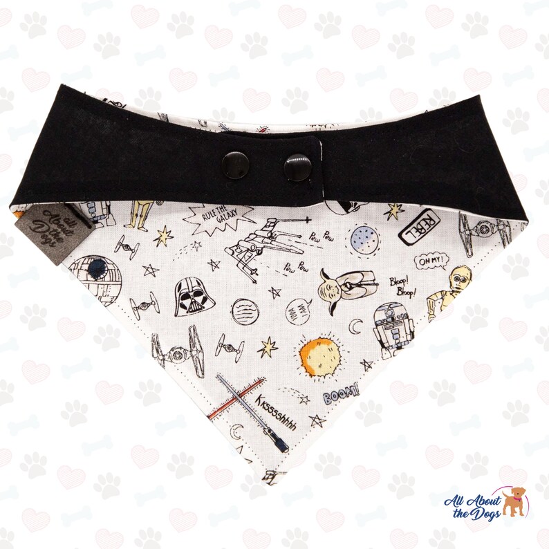 Op de afbeelding: Een witte hondenbandana met een zwarte rand en een Star Wars-thema. De bandana heeft verschillende Star Wars-personages en -symbolen, waaronder Darth Vader, R2-D2 en de Millennium Falcon. De bandana heeft een knoopsluiting.