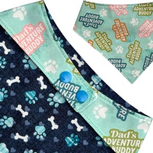 Puede incluir: Bandana para perros con fondo azul marino con patrones de huesos y huellas de patas blancas. La bandana tiene un ribete verde azulado claro con el texto "Dad's Adventure Buddy" y cierres de bot&oacute;n a presi&oacute;n azules.