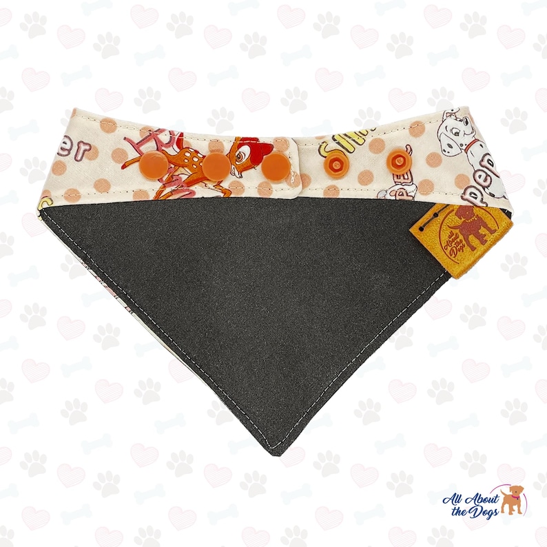 May include: A white and orange dog bandana with a black and gray backing. The bandana features a cartoon image of a deer and a Dalmatian dog. The bandana has a brown leather tag with the text "All About the Dogs" and a paw print.