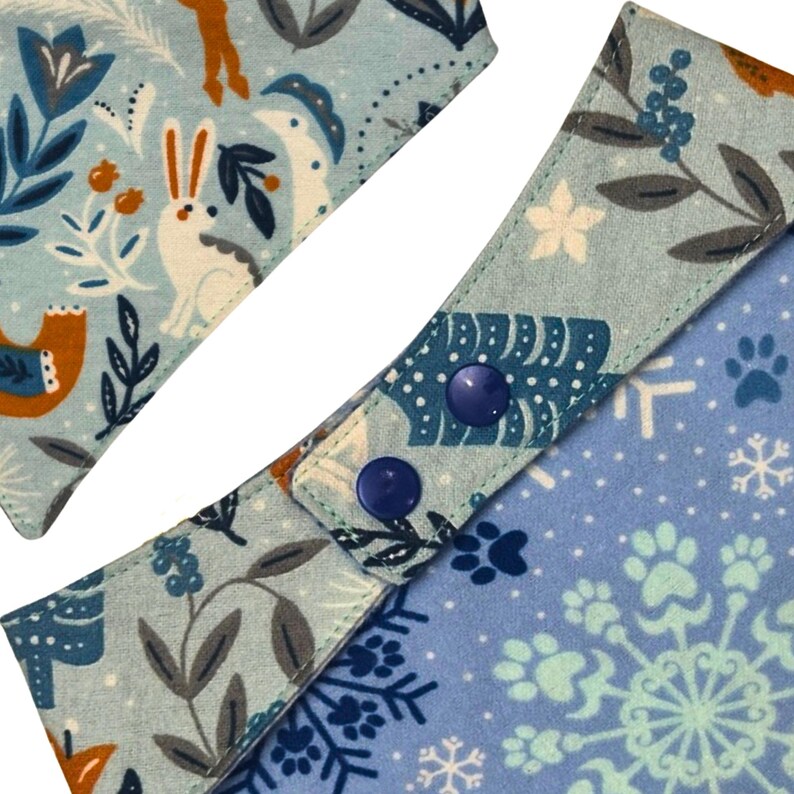 May include: Close-up of two light blue dog bandanas with winter-themed patterns. One features a white rabbit, birds, and foliage, while the other has paw prints and snowflakes. Both bandanas have blue snap closures.