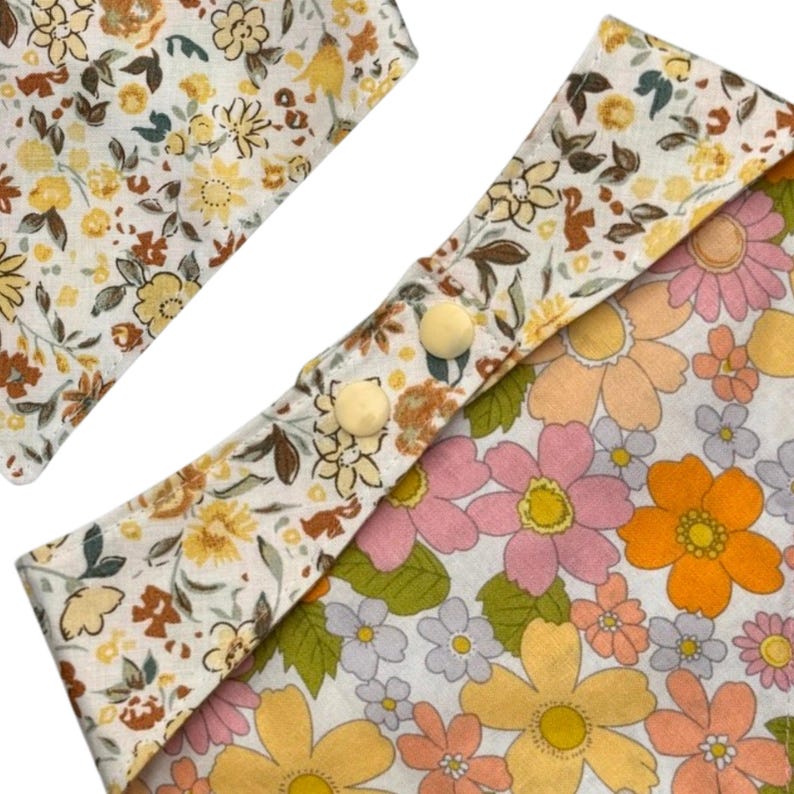 Pode incluir: Close-up de dois artigos de tecido com padr&atilde;o floral. Um &eacute; de formato quadrado com fundo branco e flores amarelas, castanhas e verdes. O outro &eacute; um formato semelhante a um babete com padr&atilde;o floral e dois bot&otilde;es creme. O segundo artigo tem fundo branco com flores rosa, laranja e amarelas.