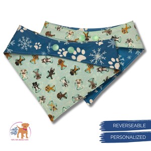 K&ouml;nnte beinhalten: Wende-Hunde-Bandana mit blauem Schneeflocken- und Pfotenabdruck-Design auf der einen Seite und einem hellblauen Muster mit Hunden auf der anderen Seite. Mit Druckkn&ouml;pfen. Die Texte "REVERSEABLE" und "PERSONALIZED" sind sichtbar.