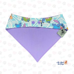 Puede incluir: Un pa&ntilde;uelo para perros con una parte superior azul claro con un estampado de personaje de dibujos animados y una parte inferior lavanda. La parte superior tiene un patr&oacute;n geom&eacute;trico y dos botones morados. El pa&ntilde;uelo es triangular y tiene una etiqueta que dice "All About the Dogs".