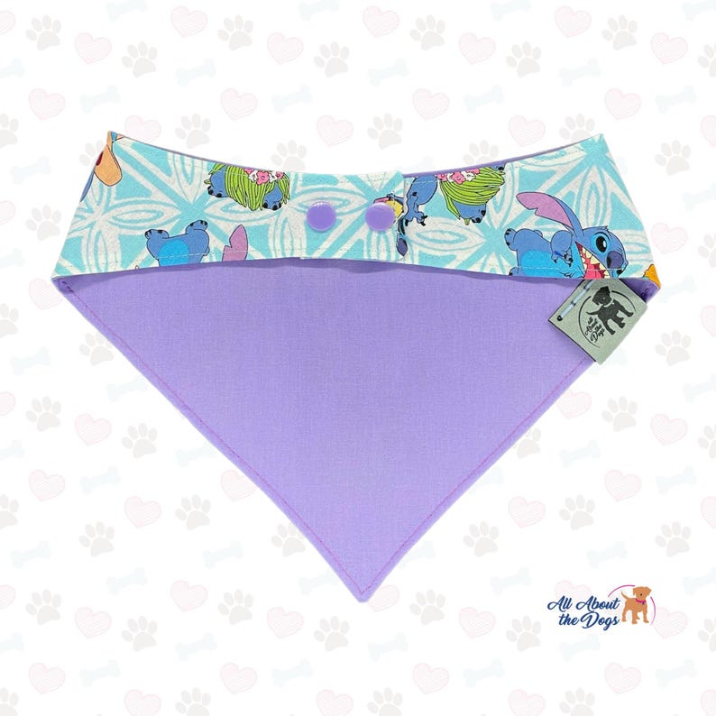 Puede incluir: Un pa&ntilde;uelo para perros con una parte superior azul claro con un estampado de personaje de dibujos animados y una parte inferior lavanda. La parte superior tiene un patr&oacute;n geom&eacute;trico y dos botones morados. El pa&ntilde;uelo es triangular y tiene una etiqueta que dice "All About the Dogs".
