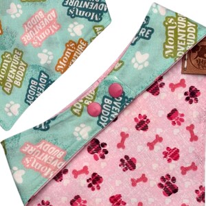 Może przedstawiać: Chusta dla psa z jasnoniebieskim i r&oacute;żowym wzorem. Jasnoniebieska sekcja zawiera napis "Mom's Adventure Buddy" i odciski łap. R&oacute;żowa sekcja ma odciski łap i kształty kości. Dwa r&oacute;żowe zatrzaski zabezpieczają chustę.