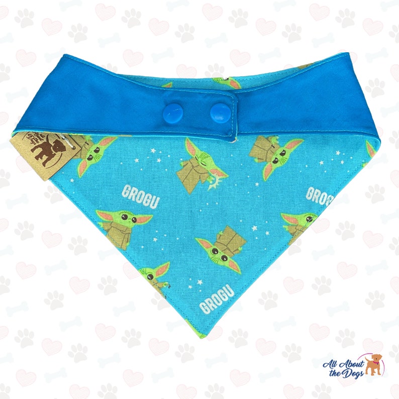 May include: A blue and green dog bandana with a repeating pattern of a green character with the word "Grogu" printed on it. The bandana has two blue buttons on the back.